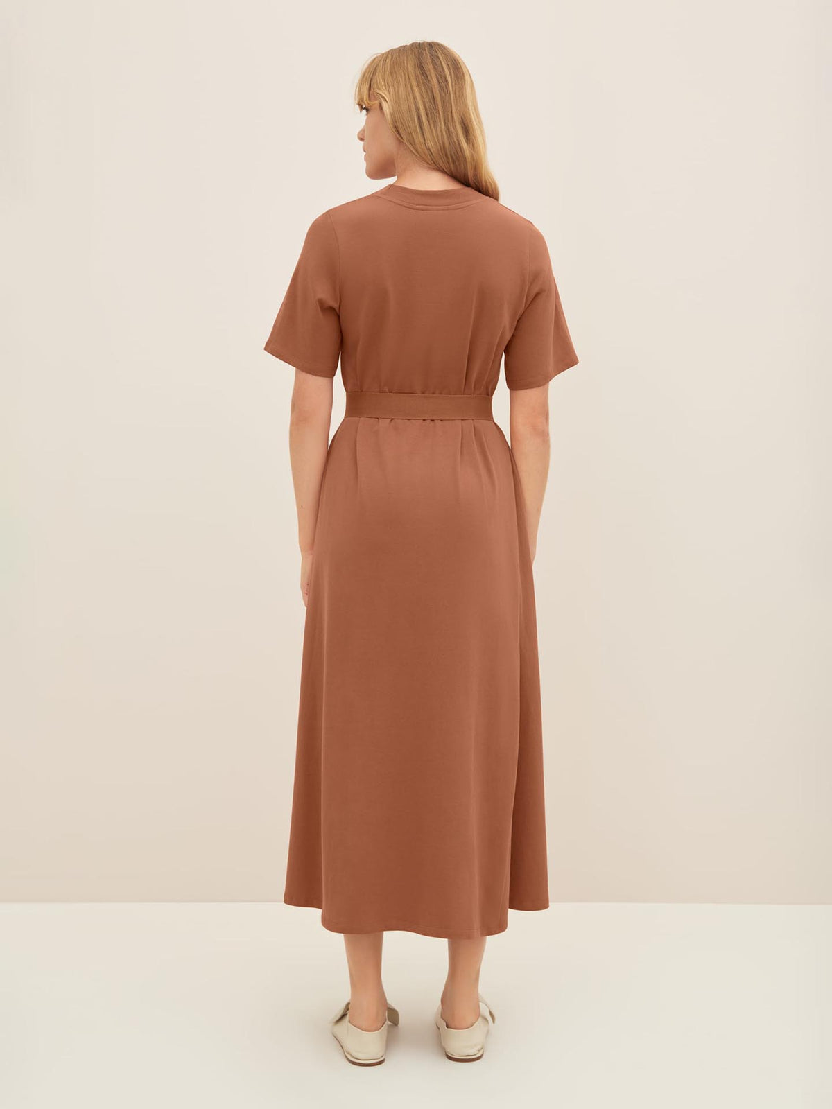 A woman with blond hair stands facing away, wearing a brown mid-length T-Shirt Swing Dress in Sienna and beige shoes against an off-white background. For those seeking the perfect standard fit, refer to the Kowtow size guide for detailed product measurements.