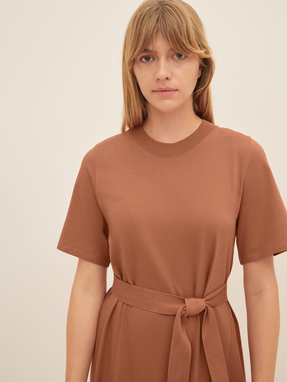 A woman with long hair wearing the Kowtow T-Shirt Swing Dress in Sienna, which features short sleeves and a tied waist, stands against a plain background. The size XS garment offers a standard fit that complements her silhouette perfectly.