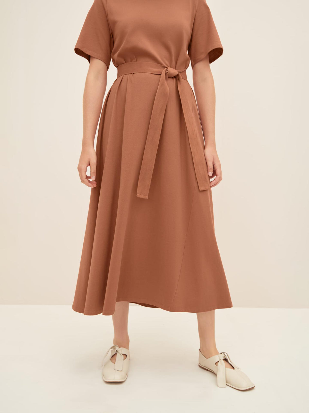 A person wearing a Kowtow T-Shirt Swing Dress in Sienna, size XS, paired with white flat shoes stands against a plain background. The brown, short-sleeved dress with a belt offers a standard fit, ideal for everyday wear.