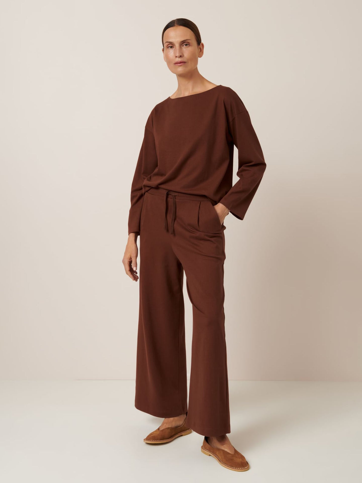 Person stands against a plain backdrop in a Kowtow ensemble: Wide Leg Pant – Sequoia, a brown long-sleeve top, and slip-on shoes styled for relaxed fit. Hands in pockets, the look is casually sophisticated. Consult the size chart for precise measurements.