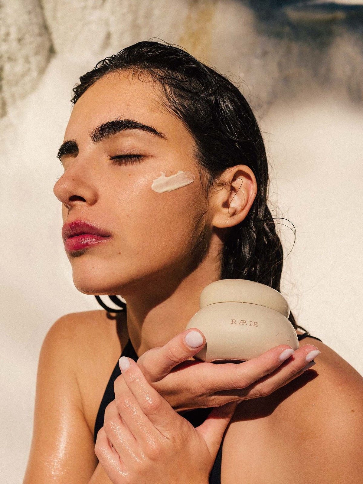 Person with wet hair holds a Cocoon Ceramide Cream container from RAAIE near their face, with a dab of hydrating moisturiser applied to their cheek. Eyes closed, they appear to be in a serene, sunny setting.