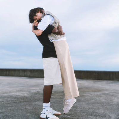 Two people embrace outdoors, with one person lifting the other off the ground. Both wear casual, light-colored clothing. The background features a cloudy sky and concrete surface.