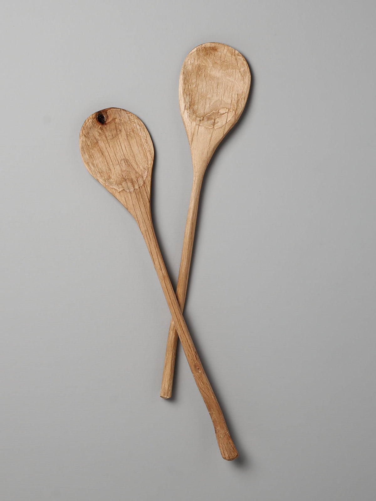 Two rustic wooden Paddle Spoons by Luke Crouch, with worn surfaces and long handles, are crossed against a plain gray background. One spoon, crafted from Australian She-Oak, features a small hole near the top.