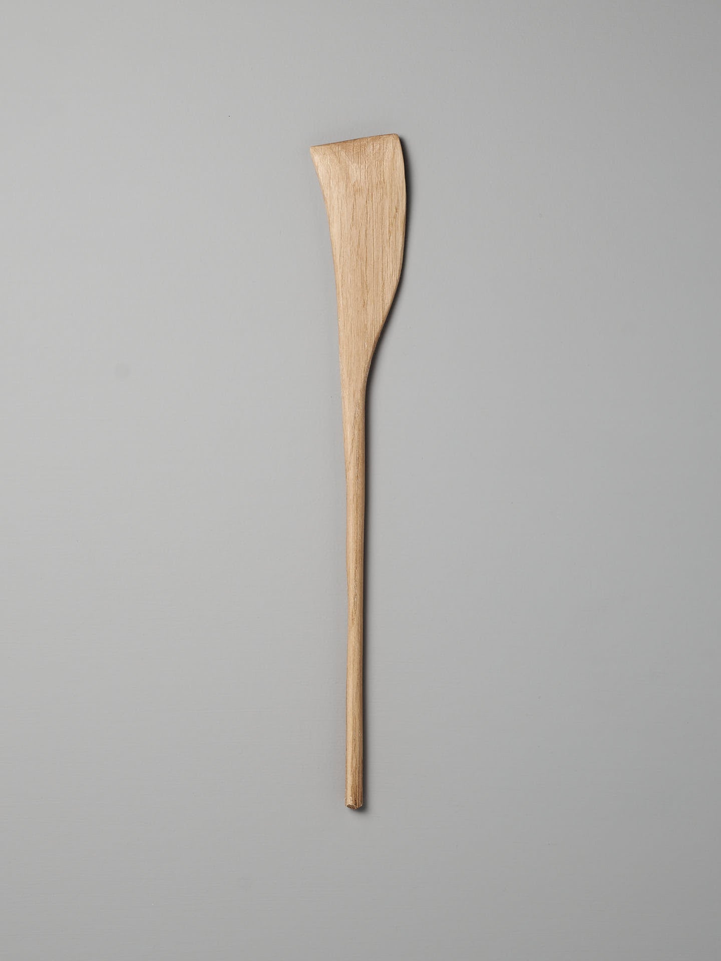 A Luke Crouch Sauté Tool, with its hand-carved wooden craftsmanship, features a slim, elongated handle and a wider, flat edge. Nestled against a grey background, it exemplifies the charm of rustic kitchen tools. Made from Australian She-Oak.