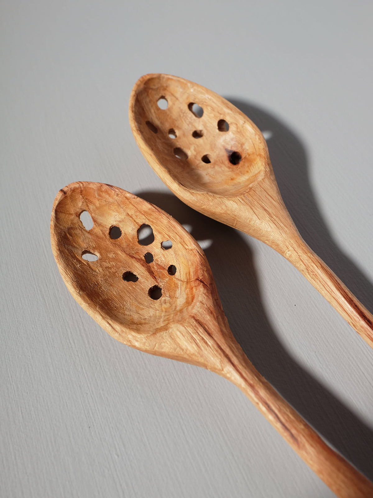 Two handcrafted Slotted Ladles from Luke Crouch, carved from Australian She-Oak using traditional techniques, each with a series of holes in their bowls, lie side by side on a smooth, light gray surface.