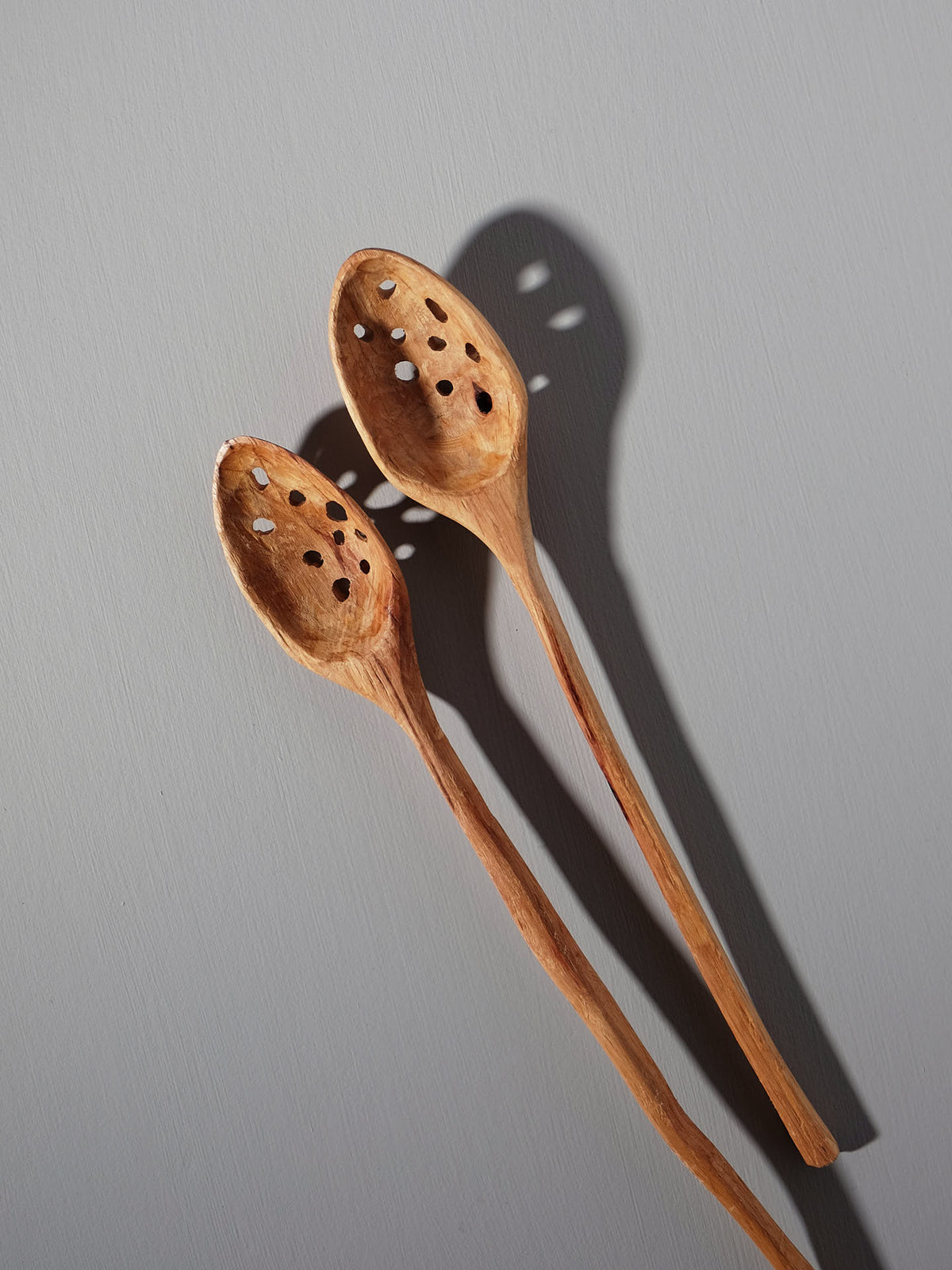 A Slotted Ladle from Luke Crouch, hand-carved from Australian She-Oak and featuring intricate perforated details, is laid against a light grey background, casting shadows to the right.