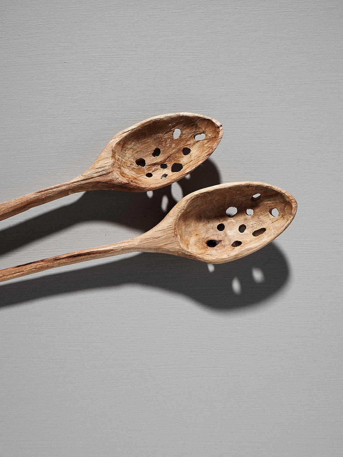 Two Slotted Ladles, hand-carved by Luke Crouch from Australian She-Oak, feature multiple holes in their wooden bowls and are placed overlapping on a light grey background.