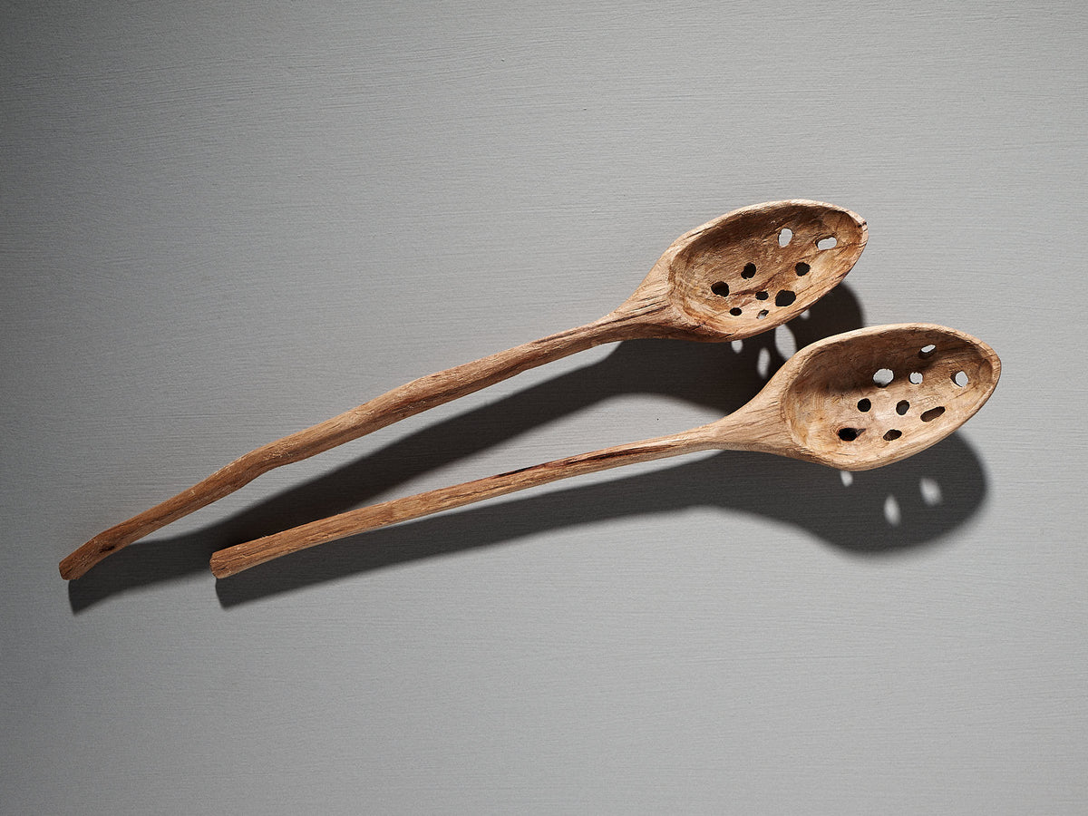 Two hand-carved kitchen utensils by Luke Crouch, including a slotted ladle crafted from Australian She-Oak using traditional techniques, lay parallel on a gray surface, casting shadows to the right.