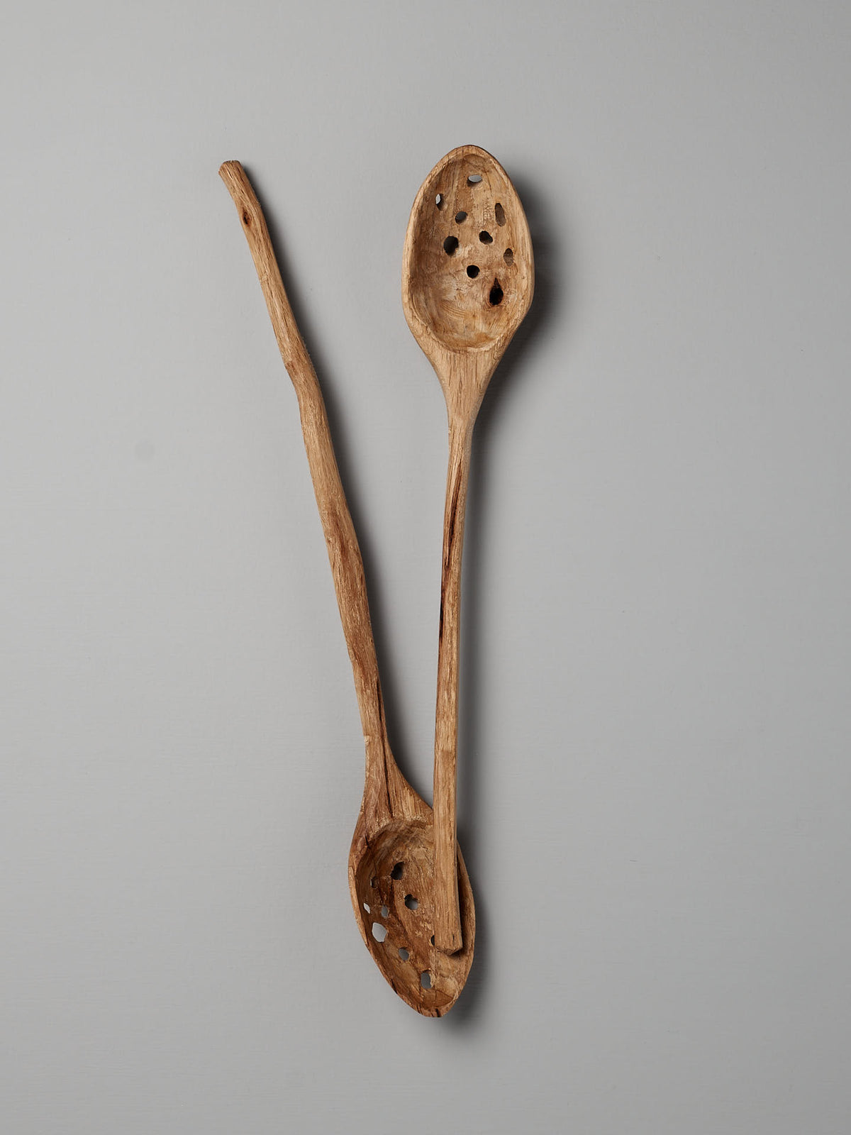 Two rustic wooden kitchen utensils from the Luke Crouch collection, featuring a hand-carved slotted ladle with drainage holes and another worn and weathered spoon, are positioned in a crisscross manner on a plain grey background.