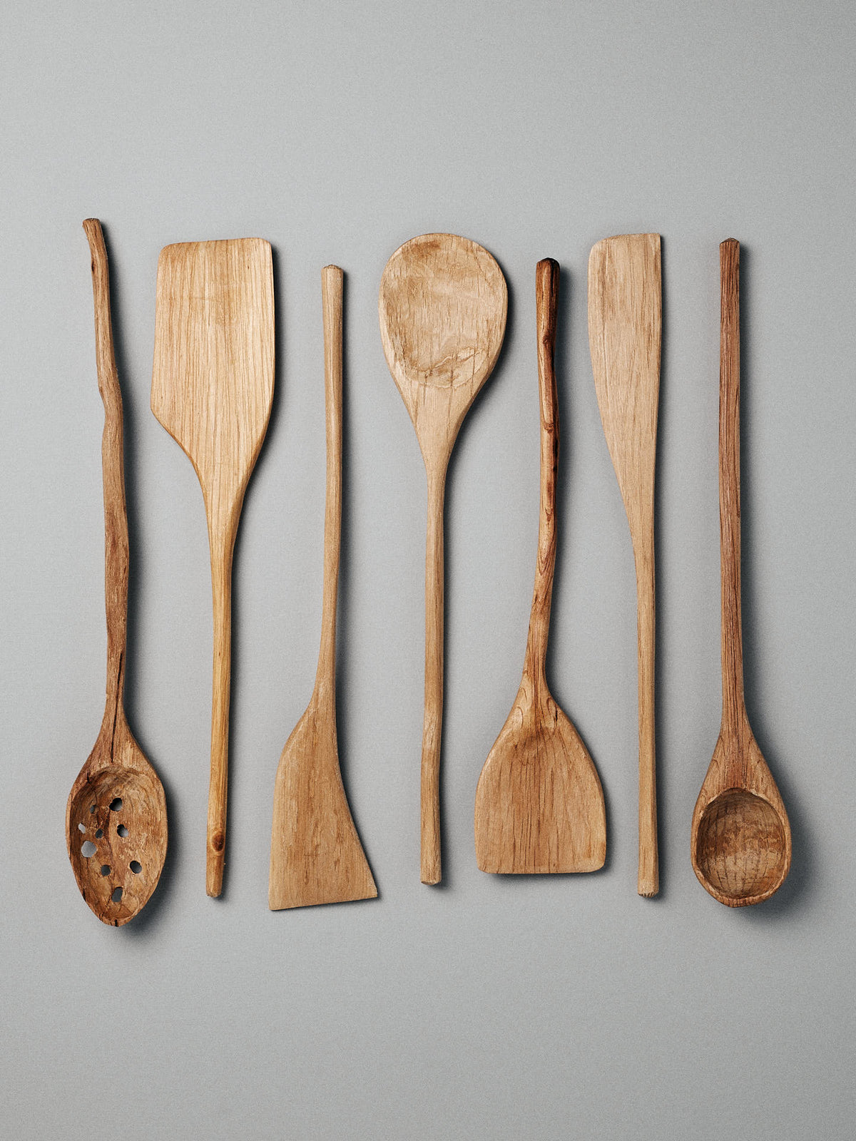 A set of seven hand-carved wooden cooking utensils by Luke Crouch, crafted from Australian She-Oak, featuring various spoons, spatulas (Product Name: Spatula), and a slotted spoon, are arranged in a row against a light gray background.