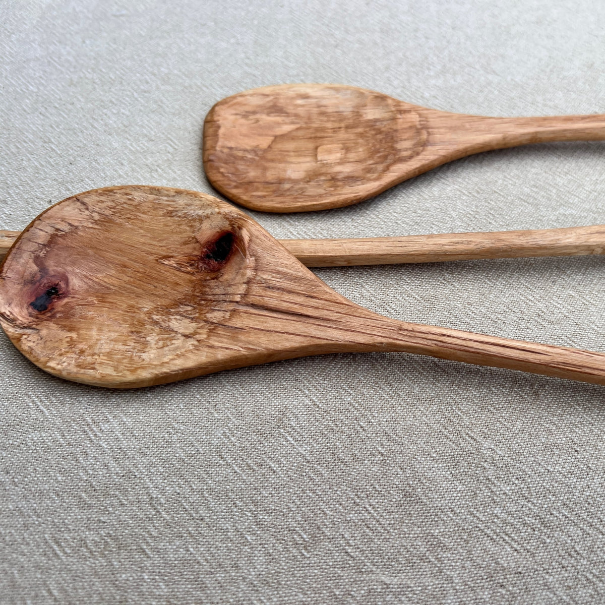 Two Paddle Spoons from Luke Crouch, hand-carved with the visible wood grain and knots of Australian She-Oak, rest elegantly on a light beige fabric surface.