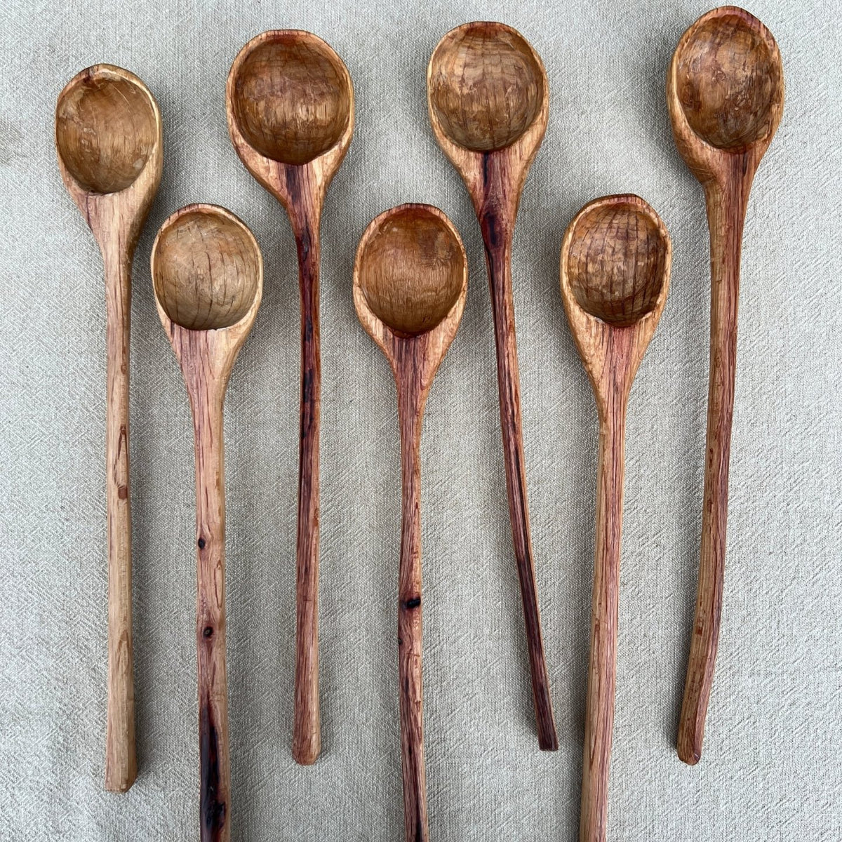 A set of seven handcrafted wooden Straight Ladles by Luke Crouch, made from Australian She-Oak, is arranged on a light fabric surface. Each hand-carved ladle features a unique, natural wood grain and a round bowl, making them distinctive kitchen accessories.