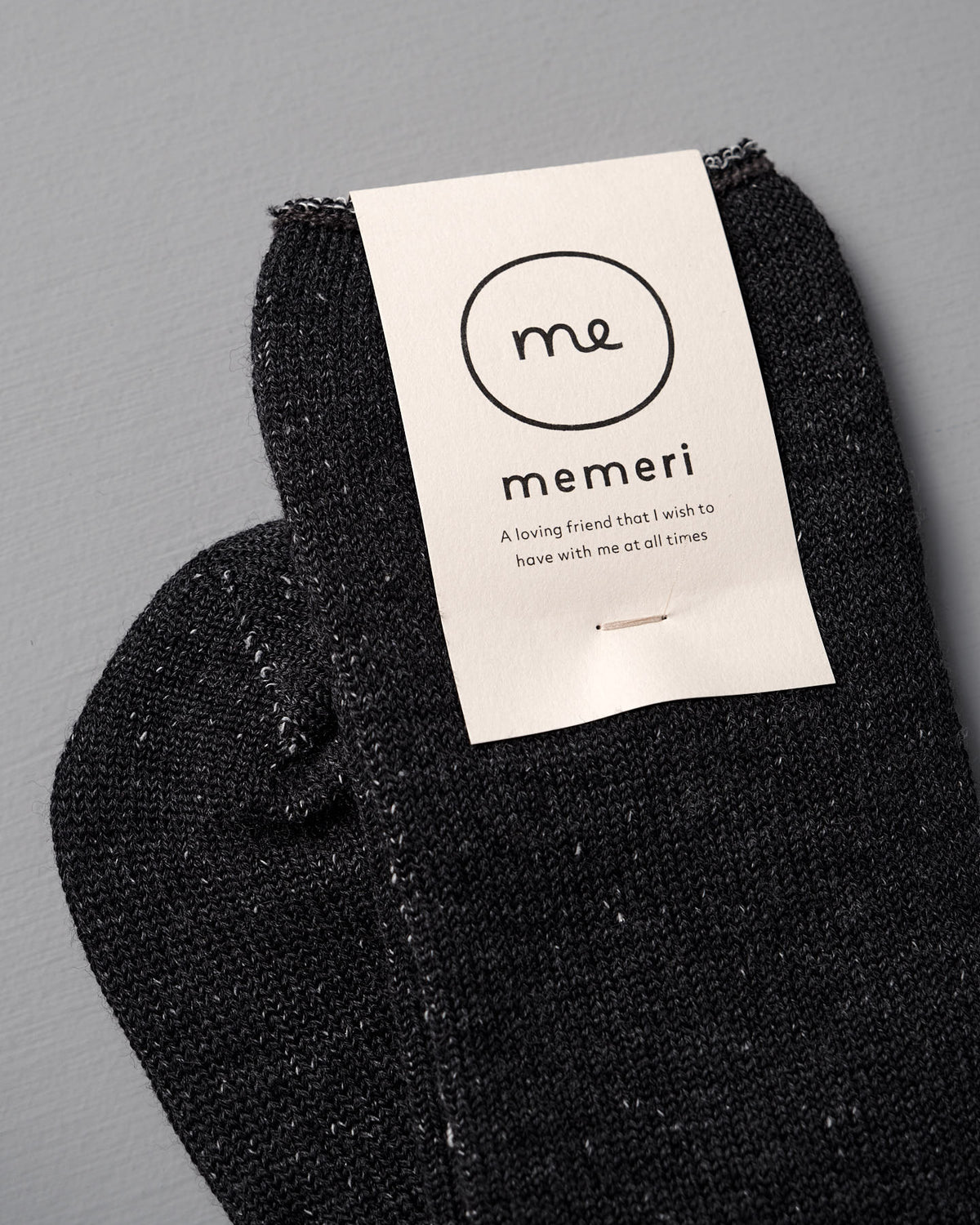 A pair of Memeri Wool Silk Socks in Charcoal by Nishiguchi Kutsushita, featuring a cream "memeri" label and short message, photographed on a gray surface.