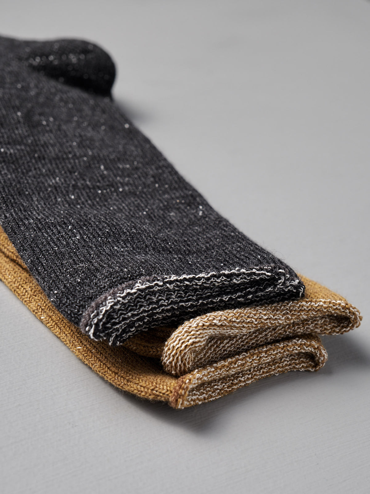 Two pairs of Nishiguchi Kutsushita Memeri Wool Silk Socks in charcoal are folded and stacked on a light gray surface.