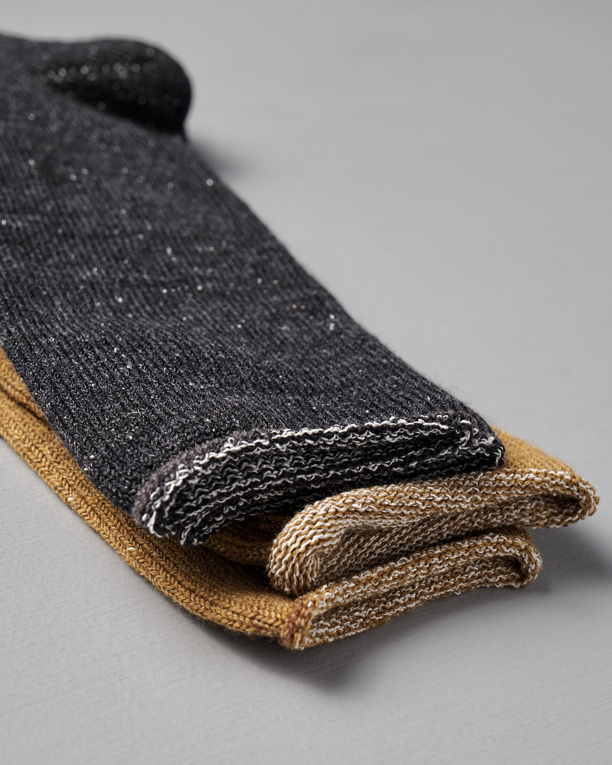 Two pairs of Nishiguchi Kutsushita Memeri Wool Silk Socks in charcoal are folded and stacked on a light gray surface.
