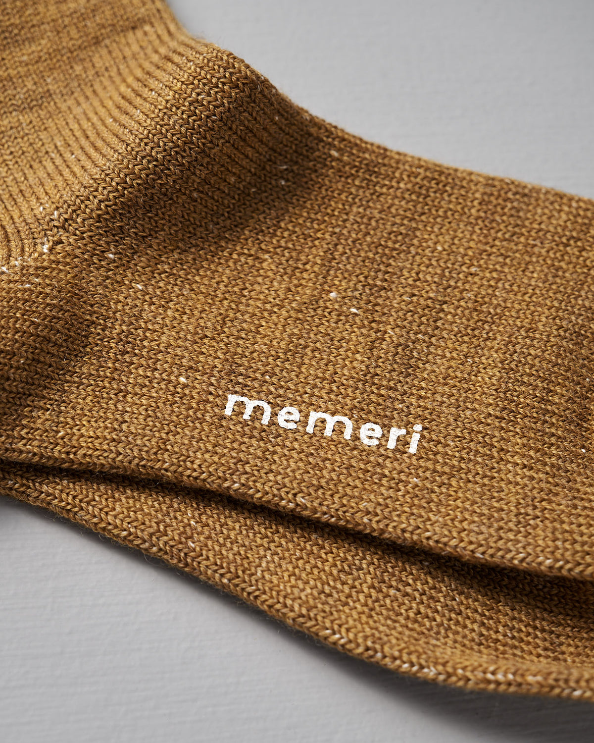 Close-up of a cozy mustard sock crafted from wool and silk by Nishiguchi Kutsushita, featuring “memeri” printed in white on the soft knit fabric.