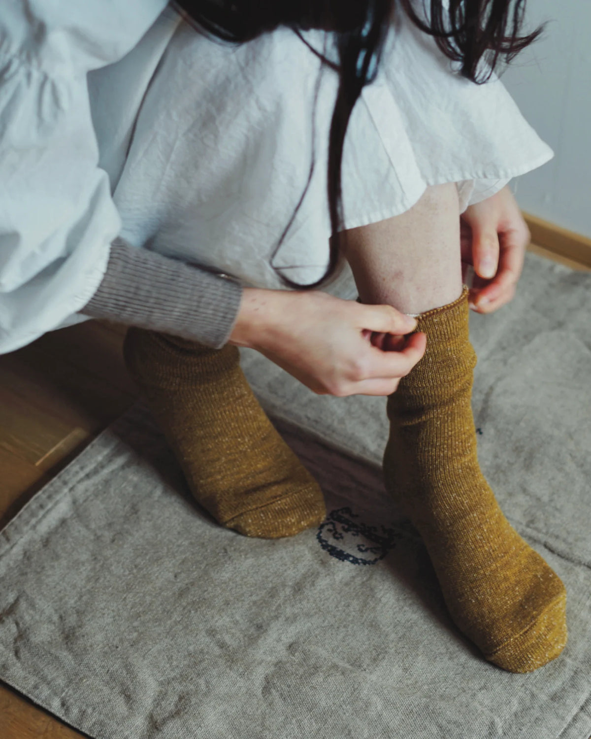 A person with long dark hair adjusts Memeri Wool Silk Socks in Mustard by Nishiguchi Kutsushita while sitting on a textured fabric mat, dressed in a white dress and gray sweater sleeves.