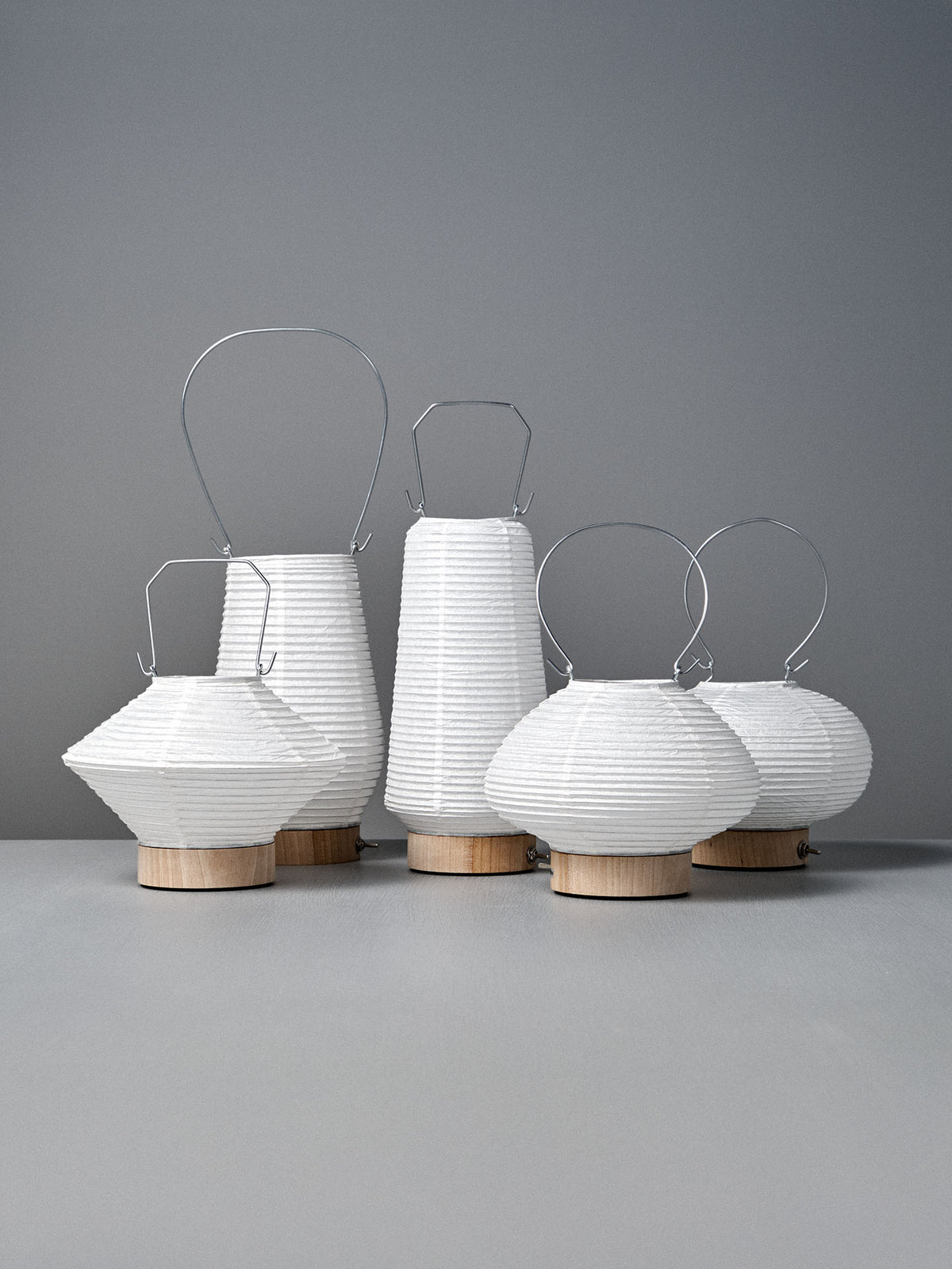 Six Washi Paper LED Lanterns – Maru, designed by Japanese product designer Rina Ono, feature white paper shades, wooden bases, and metal handles. They are arranged on a gray surface against a gray background.