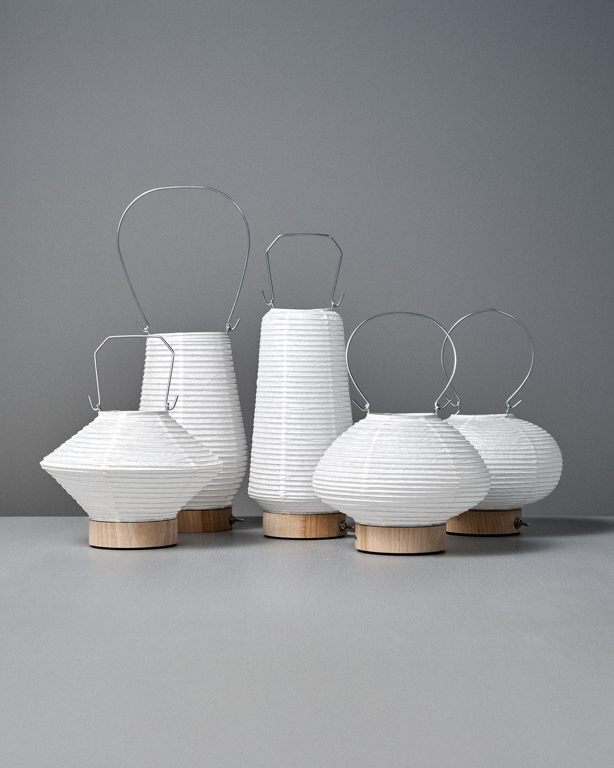 Six Washi Paper LED Lanterns – Maru, designed by Japanese product designer Rina Ono, feature white paper shades, wooden bases, and metal handles. They are arranged on a gray surface against a gray background.