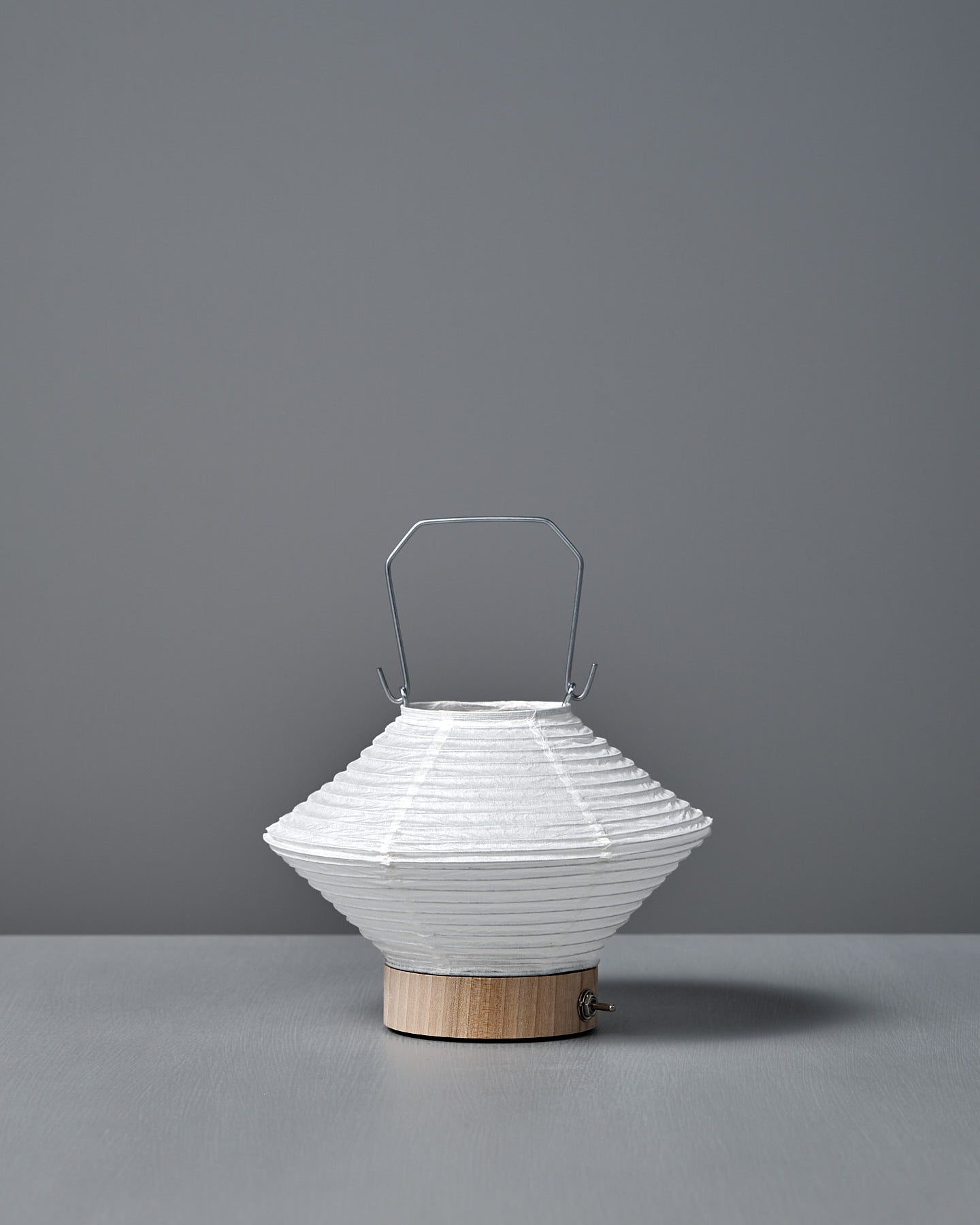 The Washi Paper LED Lantern – Hishi, featuring a white paper shade, metal handle, and wooden base by Japanese designer Rina Ono, sits on a gray surface with a gray background.