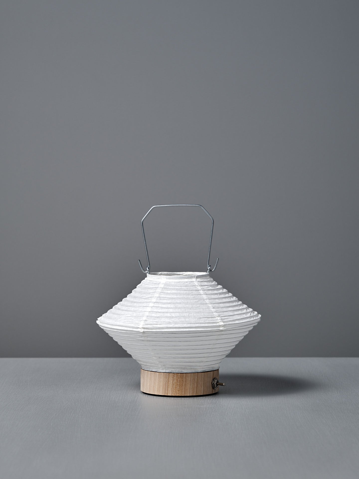 The Washi Paper LED Lantern – Hishi, featuring a white paper shade, metal handle, and wooden base by Japanese designer Rina Ono, sits on a gray surface with a gray background.