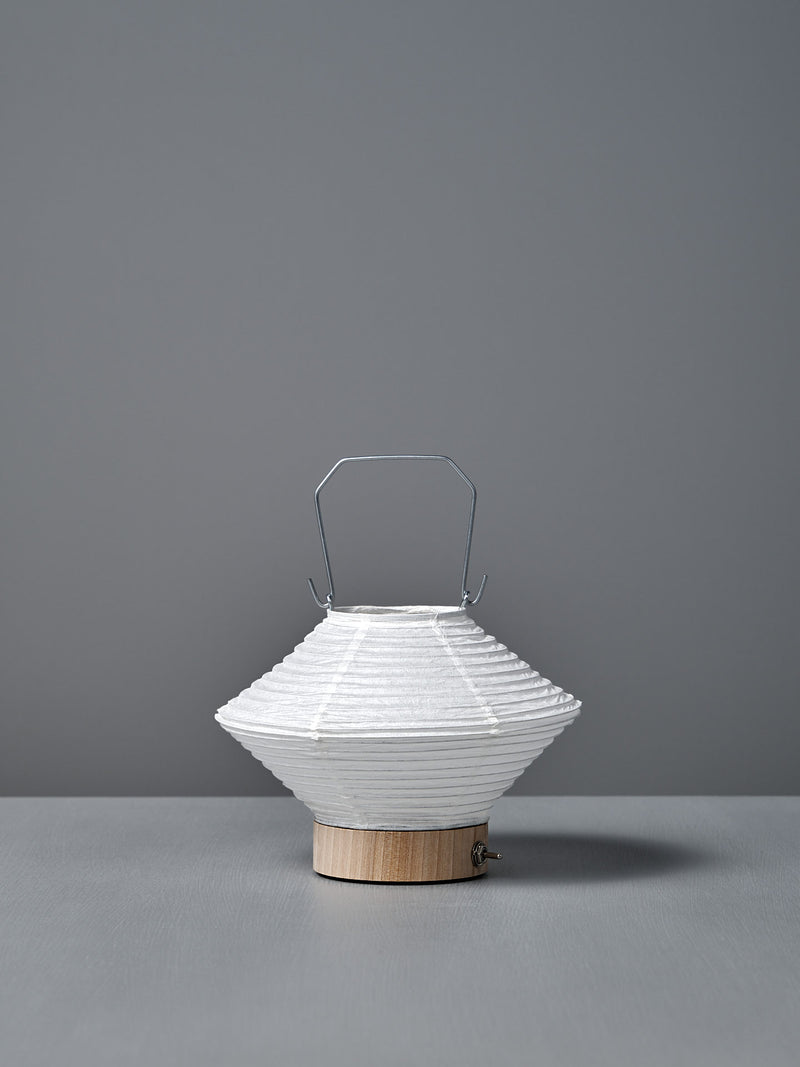 The Washi Paper LED Lantern – Hishi, featuring a white paper shade, metal handle, and wooden base by Japanese designer Rina Ono, sits on a gray surface with a gray background.