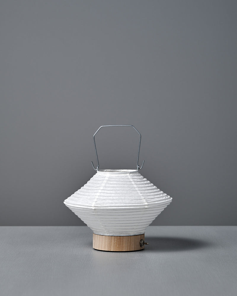 The Washi Paper LED Lantern – Hishi, featuring a white paper shade, metal handle, and wooden base by Japanese designer Rina Ono, sits on a gray surface with a gray background.