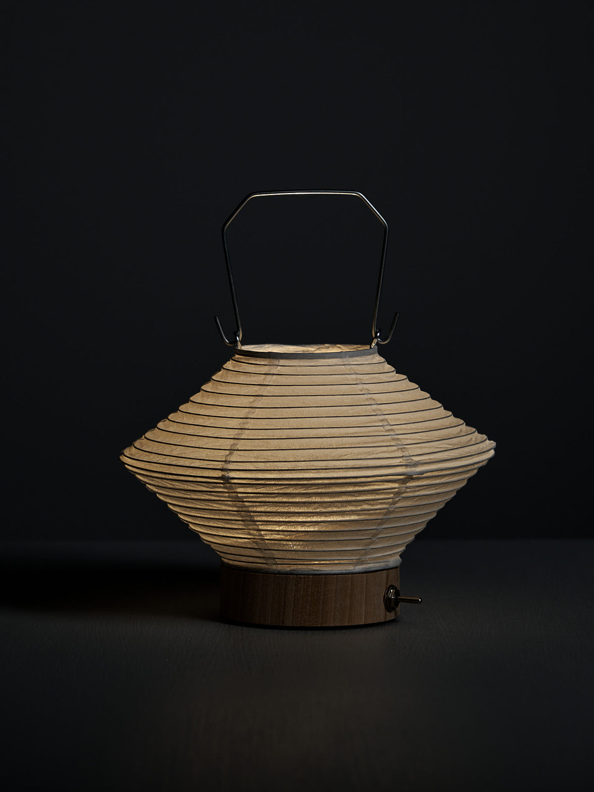 The Washi Paper LED Lantern – Hishi, designed by Rina Ono with a wooden base and metal handle, glows softly against a dark background.