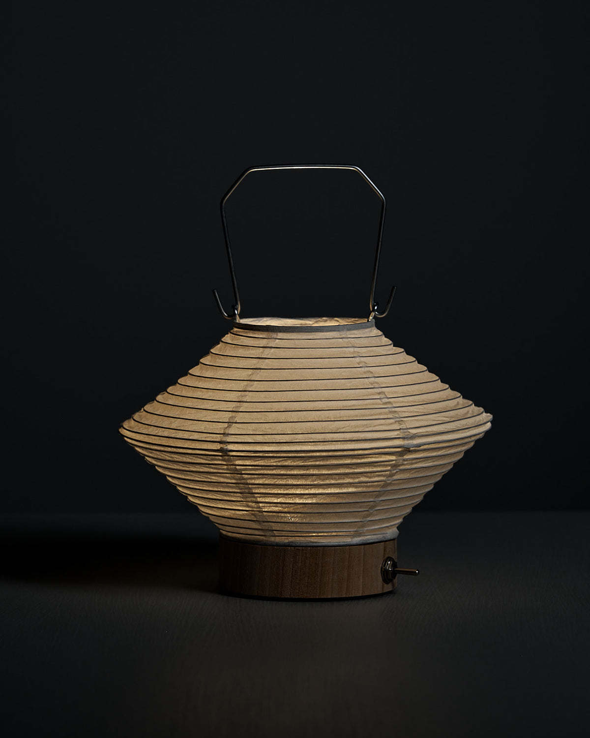 The Washi Paper LED Lantern – Hishi, designed by Rina Ono with a wooden base and metal handle, glows softly against a dark background.