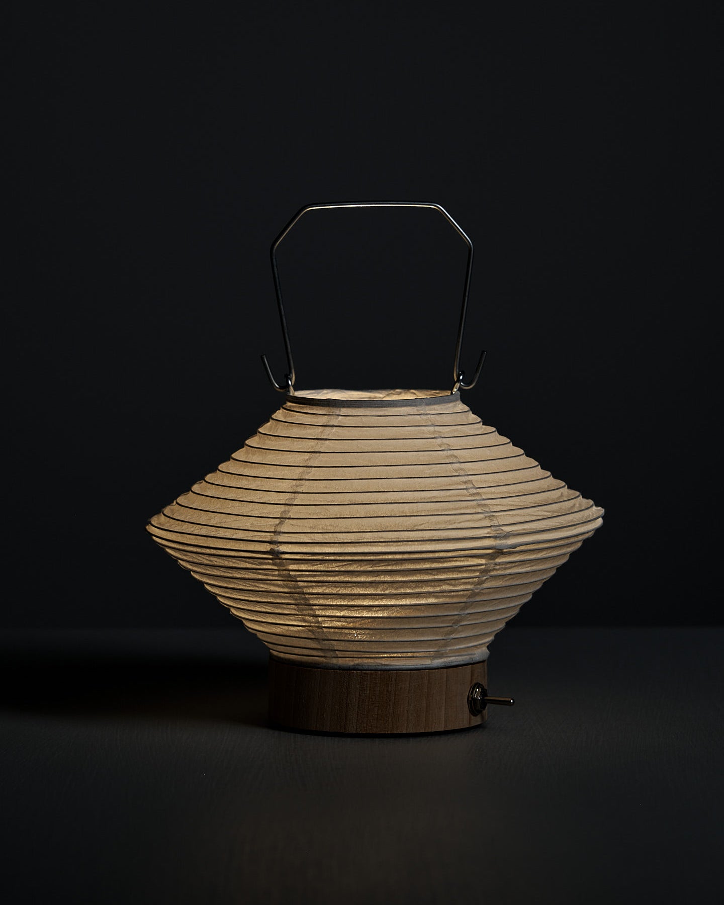 The Washi Paper LED Lantern – Hishi, featuring a white paper shade, metal handle, and wooden base by Japanese designer Rina Ono, sits on a gray surface with a gray background.