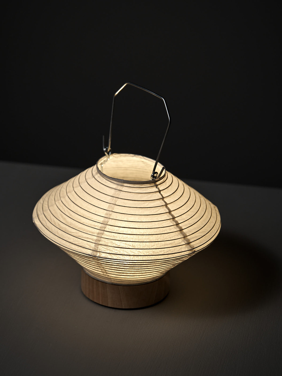 The Washi Paper LED Lantern – Hishi, designed by Rina Ono, features concentric ridges, a metal handle, and a wooden base, elegantly displayed on a dark surface.