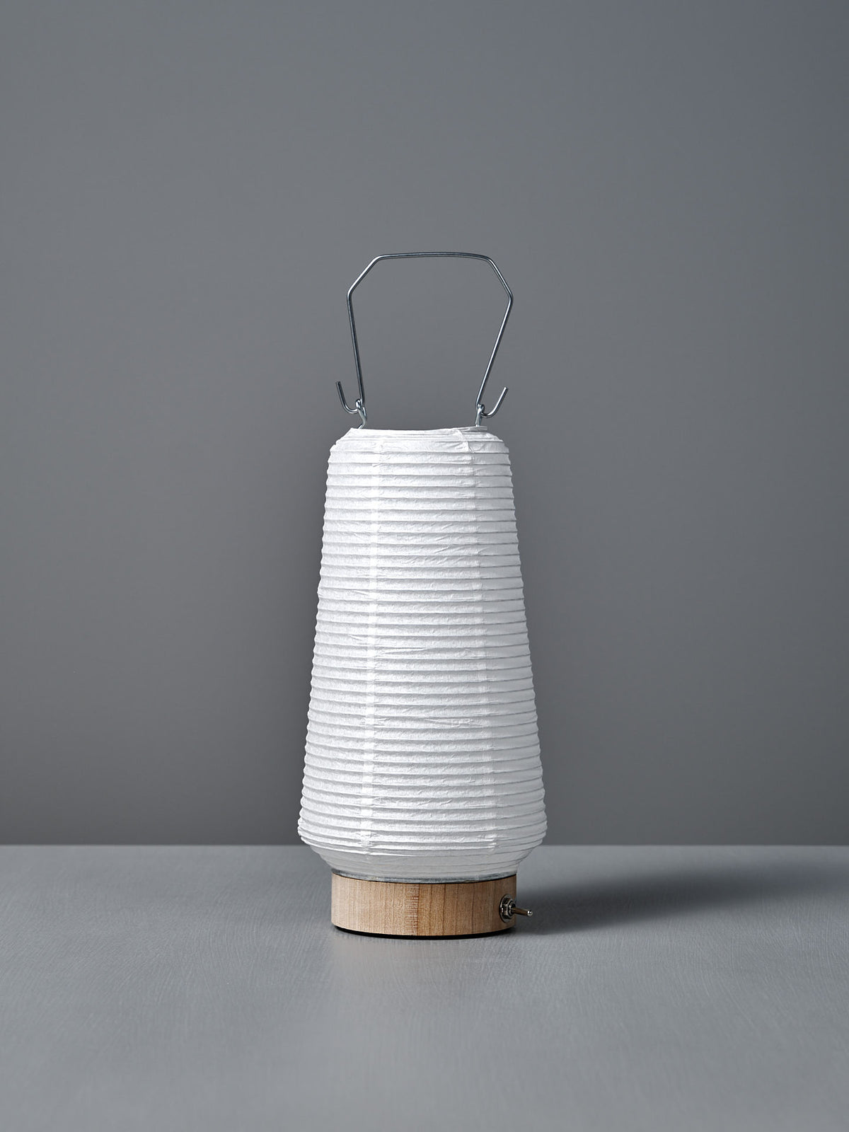 The Washi Paper LED Lantern – Itomaki, designed by Rina Ono, features a white washi paper shade, wooden base, and metal handle. Shown on a gray surface with a gray background.