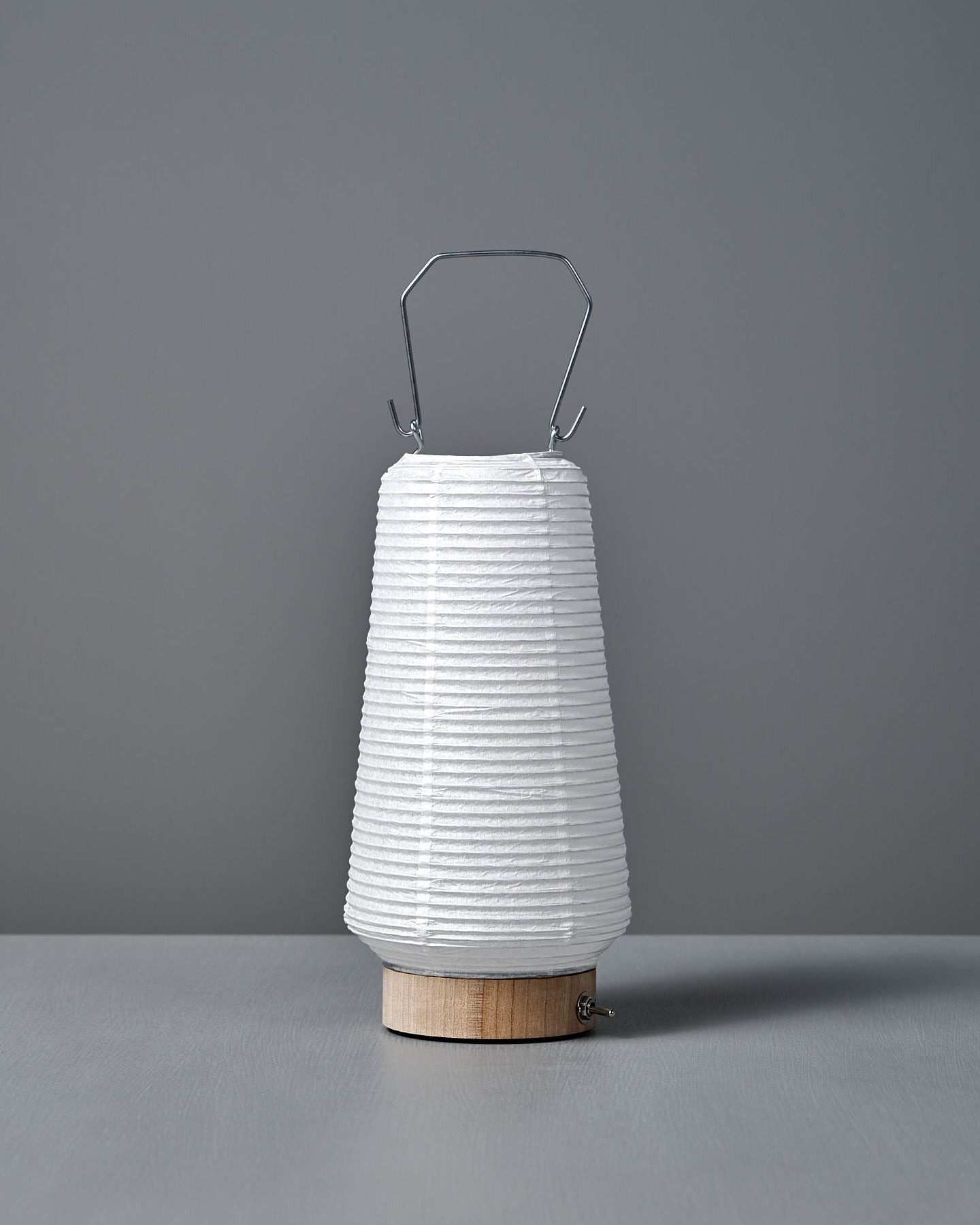The Washi Paper LED Lantern – Itomaki, designed by Rina Ono, features a white washi paper shade, wooden base, and metal handle. Shown on a gray surface with a gray background.