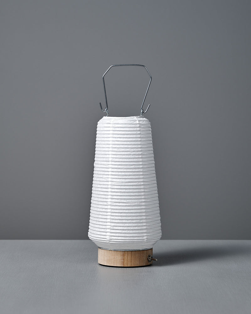 The Washi Paper LED Lantern – Itomaki, designed by Rina Ono, features a white washi paper shade, wooden base, and metal handle. Shown on a gray surface with a gray background.