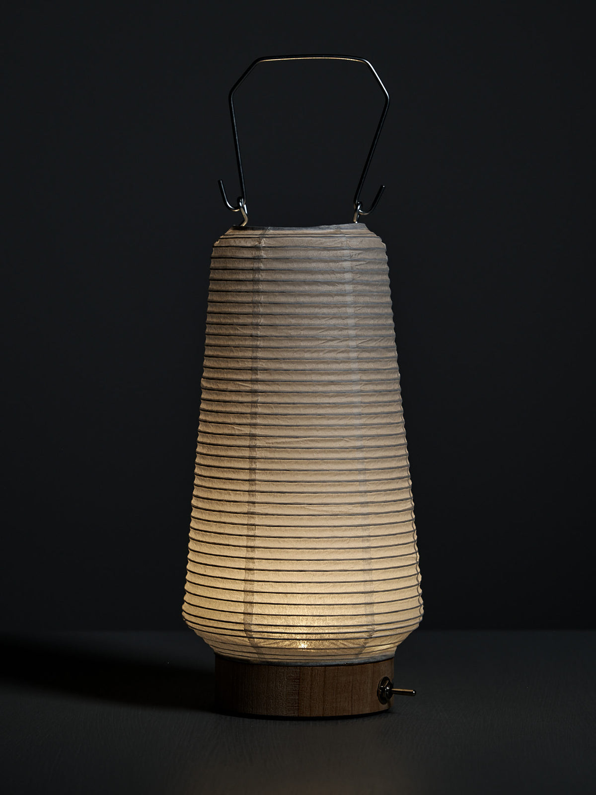 The Washi Paper LED Lantern – Itomaki, designed by Rina Ono, features a ribbed washi shade, wooden base, and metal handle, and is set against a dark background.