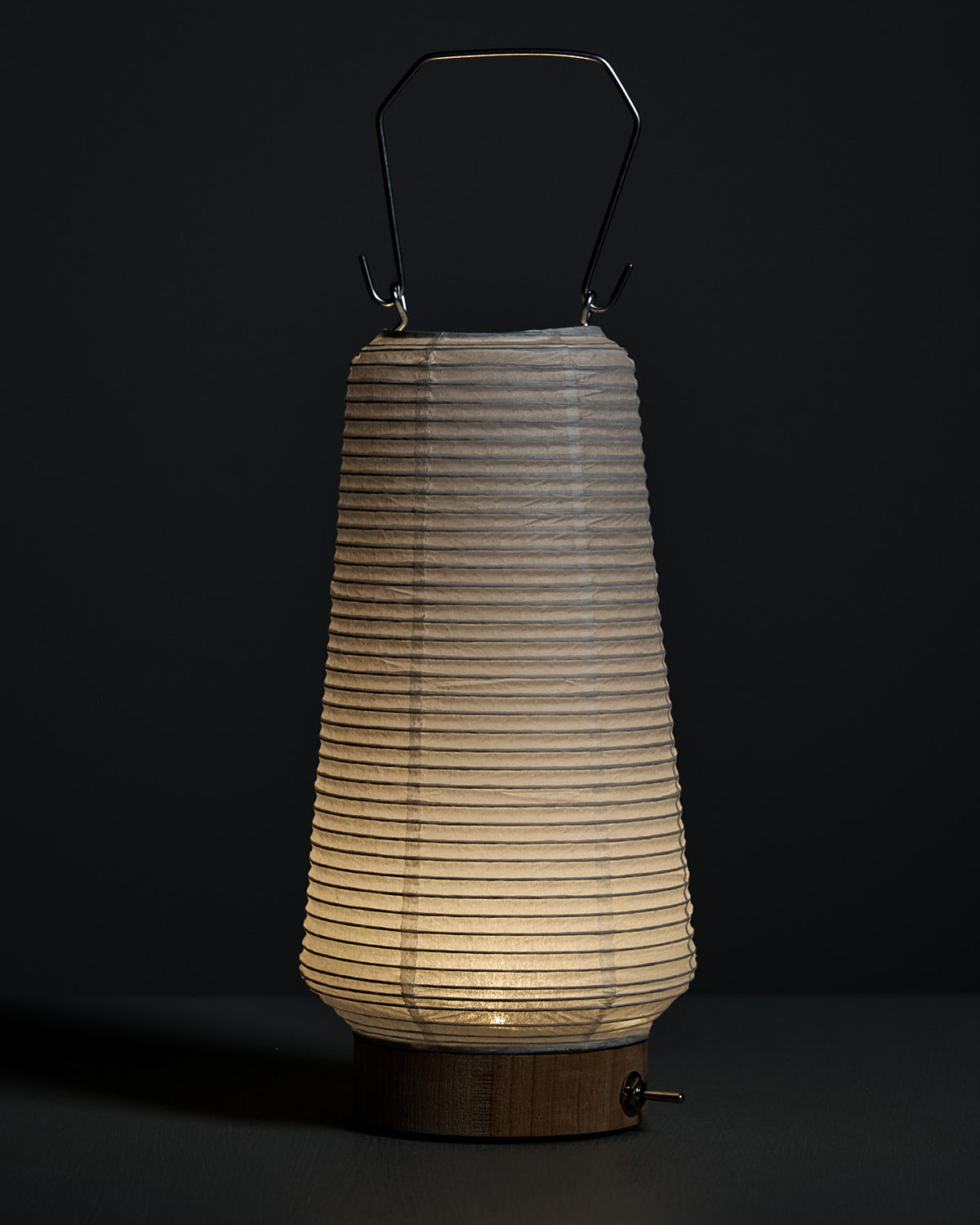The Washi Paper LED Lantern – Itomaki, designed by Rina Ono, features a ribbed washi shade, wooden base, and metal handle, and is set against a dark background.