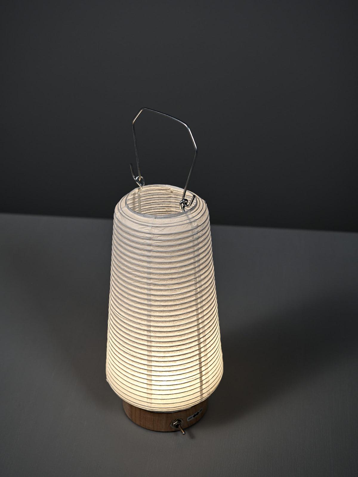 The Washi Paper LED Lantern – Itomaki, designed by Rina Ono of Rinao design studio, with a metal handle, is illuminated and placed on a gray surface against a dark gray background.