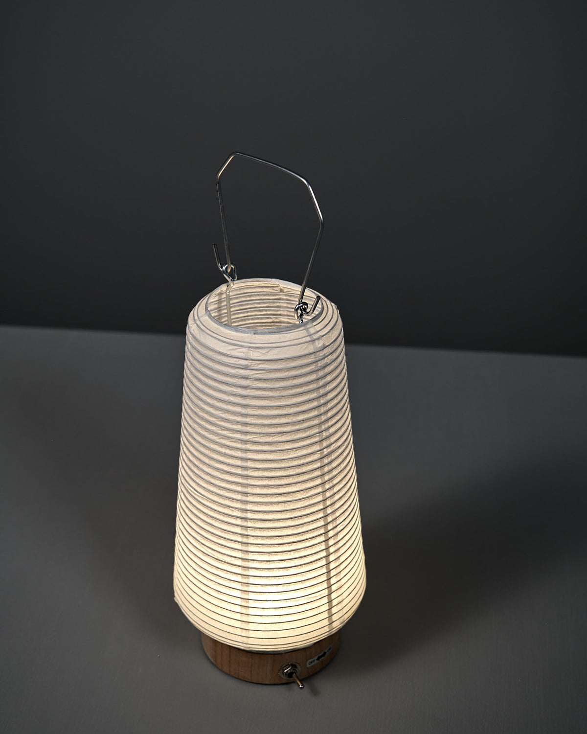 The Washi Paper LED Lantern – Itomaki, designed by Rina Ono of Rinao design studio, with a metal handle, is illuminated and placed on a gray surface against a dark gray background.