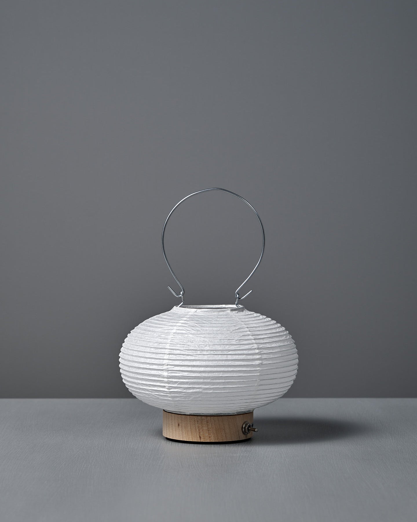 The Washi Paper LED Lantern – Maru, designed by Rina Ono, is a small, round white lantern with a metal handle and a light wooden base, displayed against a plain gray background.