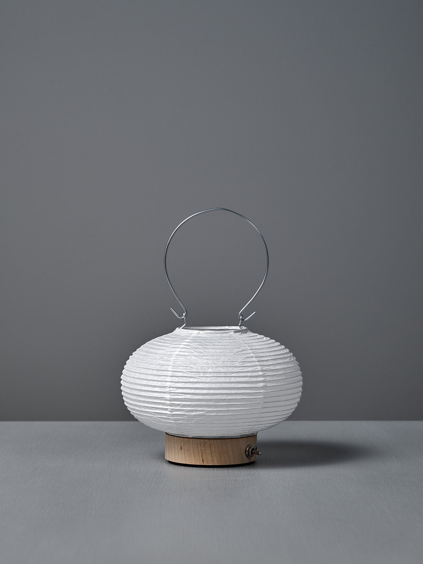 The Washi Paper LED Lantern – Maru, designed by Rina Ono, is a small, round white lantern with a metal handle and a light wooden base, displayed against a plain gray background.