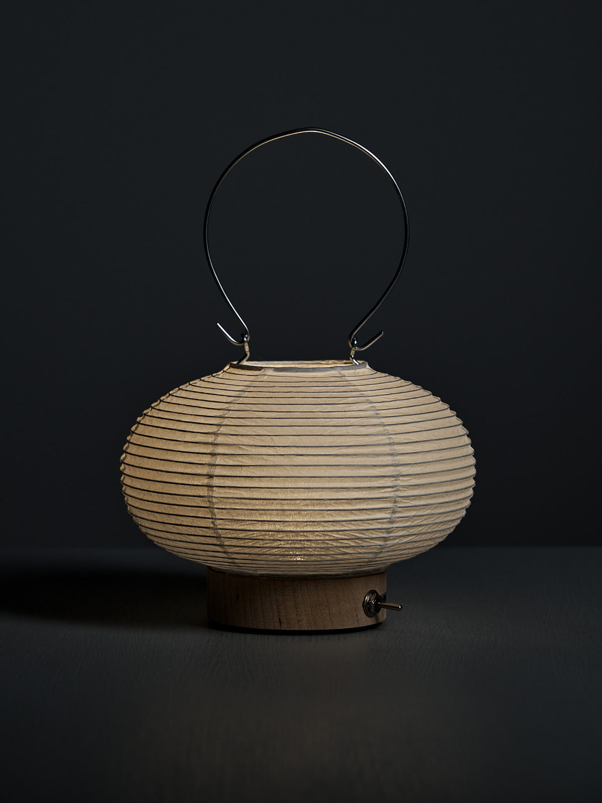The Washi Paper LED Lantern – Maru, designed by Rina Ono, features a ribbed round shade, wooden base, and metal handle. It is displayed against a dark background.