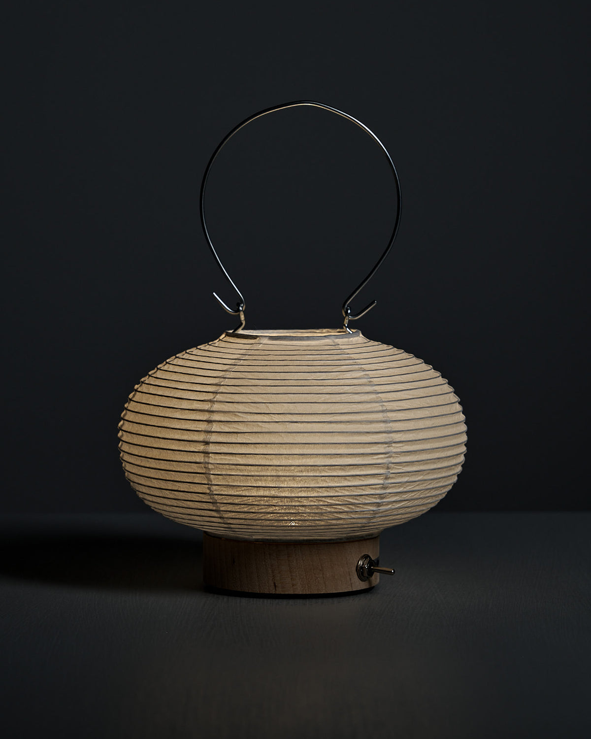 The Washi Paper LED Lantern – Maru, designed by Rina Ono, features a ribbed round shade, wooden base, and metal handle. It is displayed against a dark background.