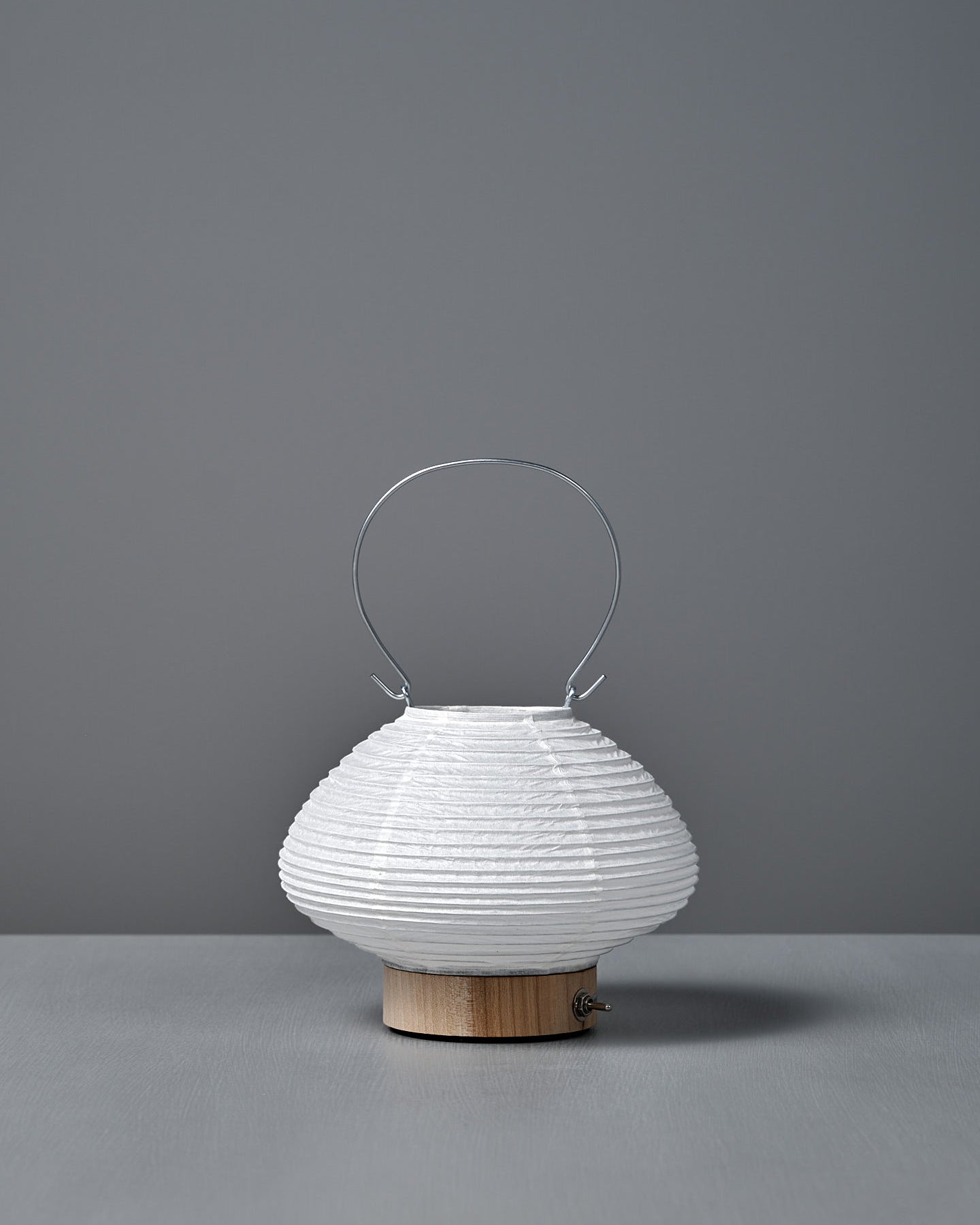 The Washi Paper LED Lantern – Musubi, designed by Rina Ono, features a small round white shape with a wooden base and thin metal handle, and sits on a grey surface against a plain grey background.