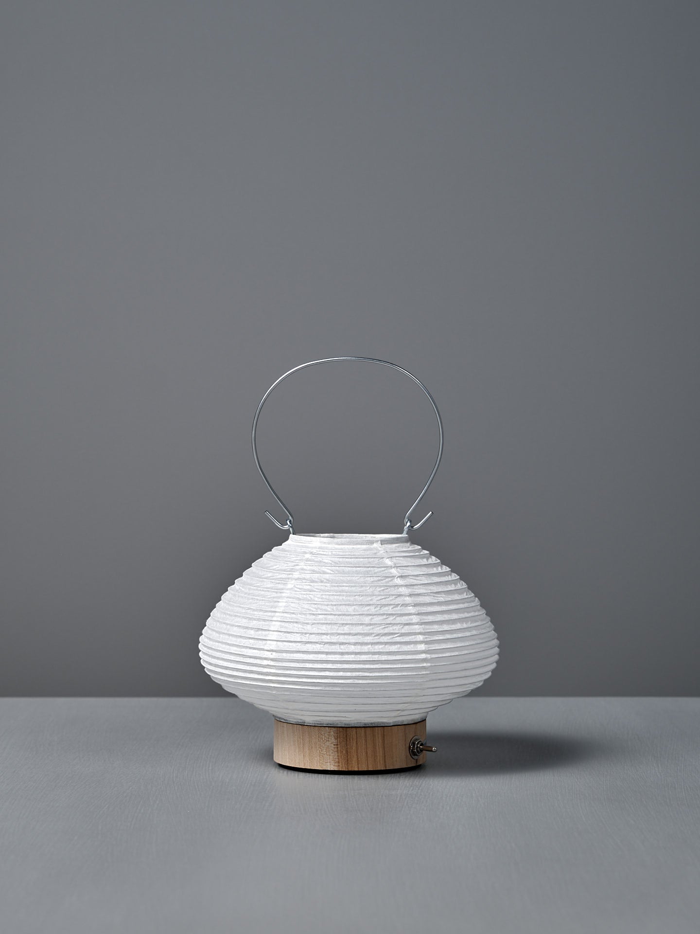 The Washi Paper LED Lantern – Musubi, designed by Rina Ono, features a small round white shape with a wooden base and thin metal handle, and sits on a grey surface against a plain grey background.