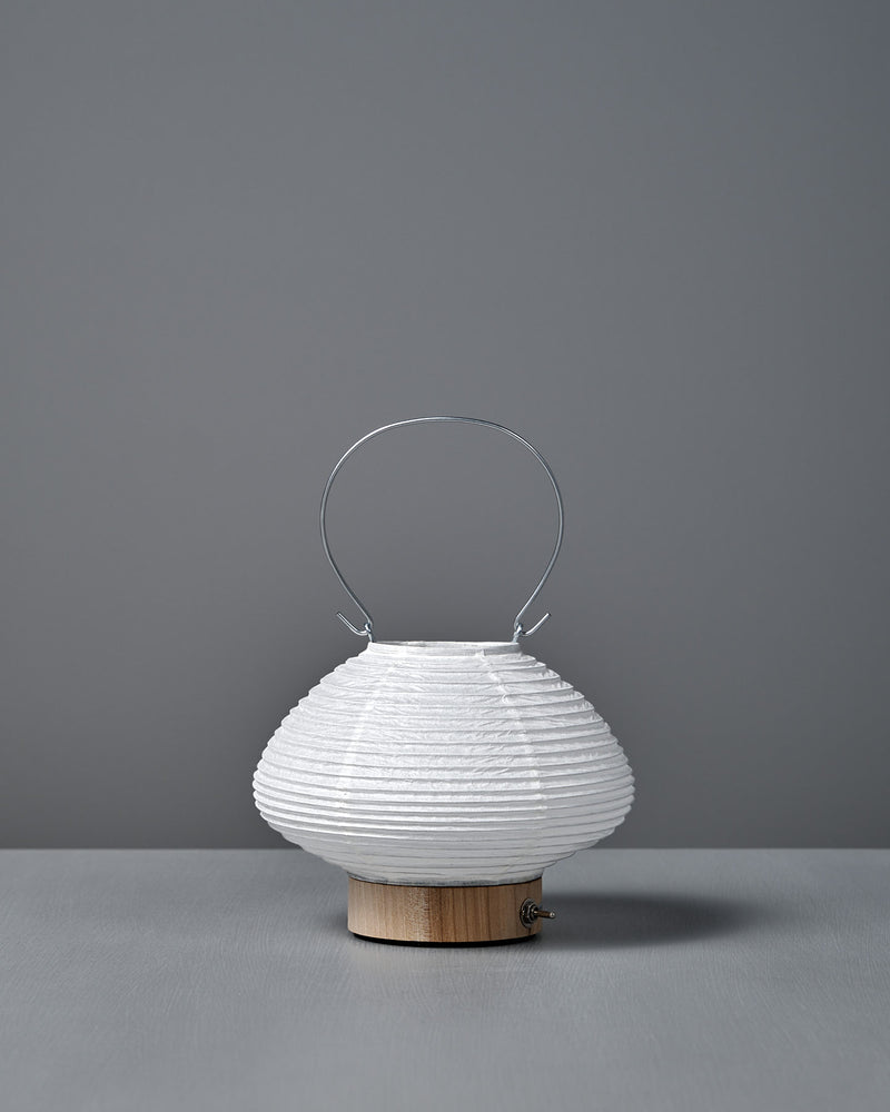 The Washi Paper LED Lantern – Musubi, designed by Rina Ono, features a small round white shape with a wooden base and thin metal handle, and sits on a grey surface against a plain grey background.