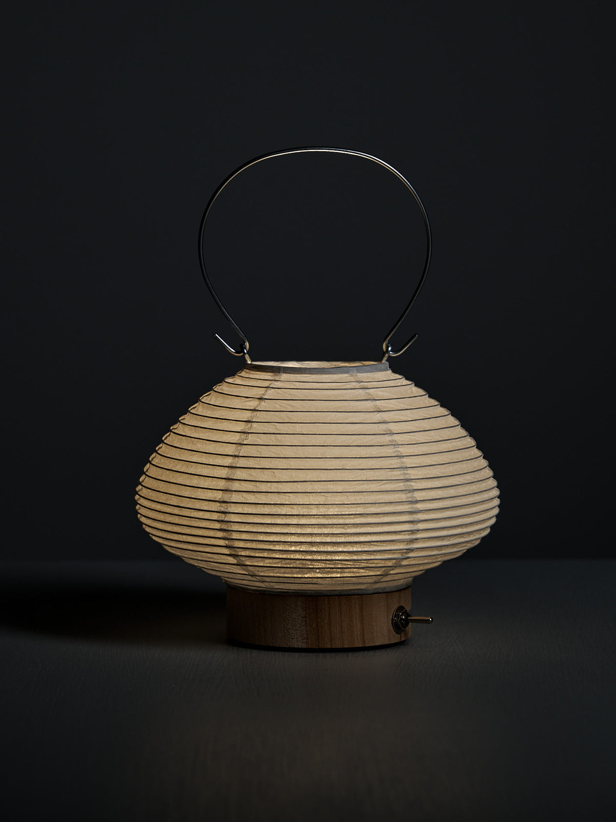 The Washi Paper LED Lantern – Musubi, featuring a rounded shape, wooden base, and metal handle by Japanese designer Rina Ono, is displayed against a dark background.