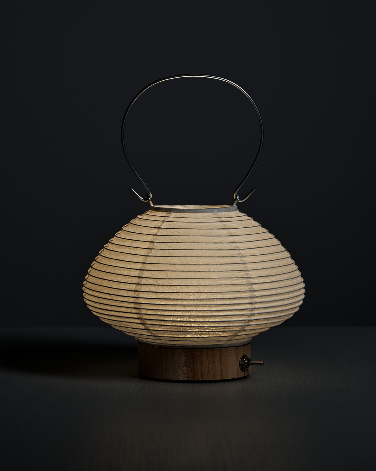The Washi Paper LED Lantern – Musubi, featuring a rounded shape, wooden base, and metal handle by Japanese designer Rina Ono, is displayed against a dark background.