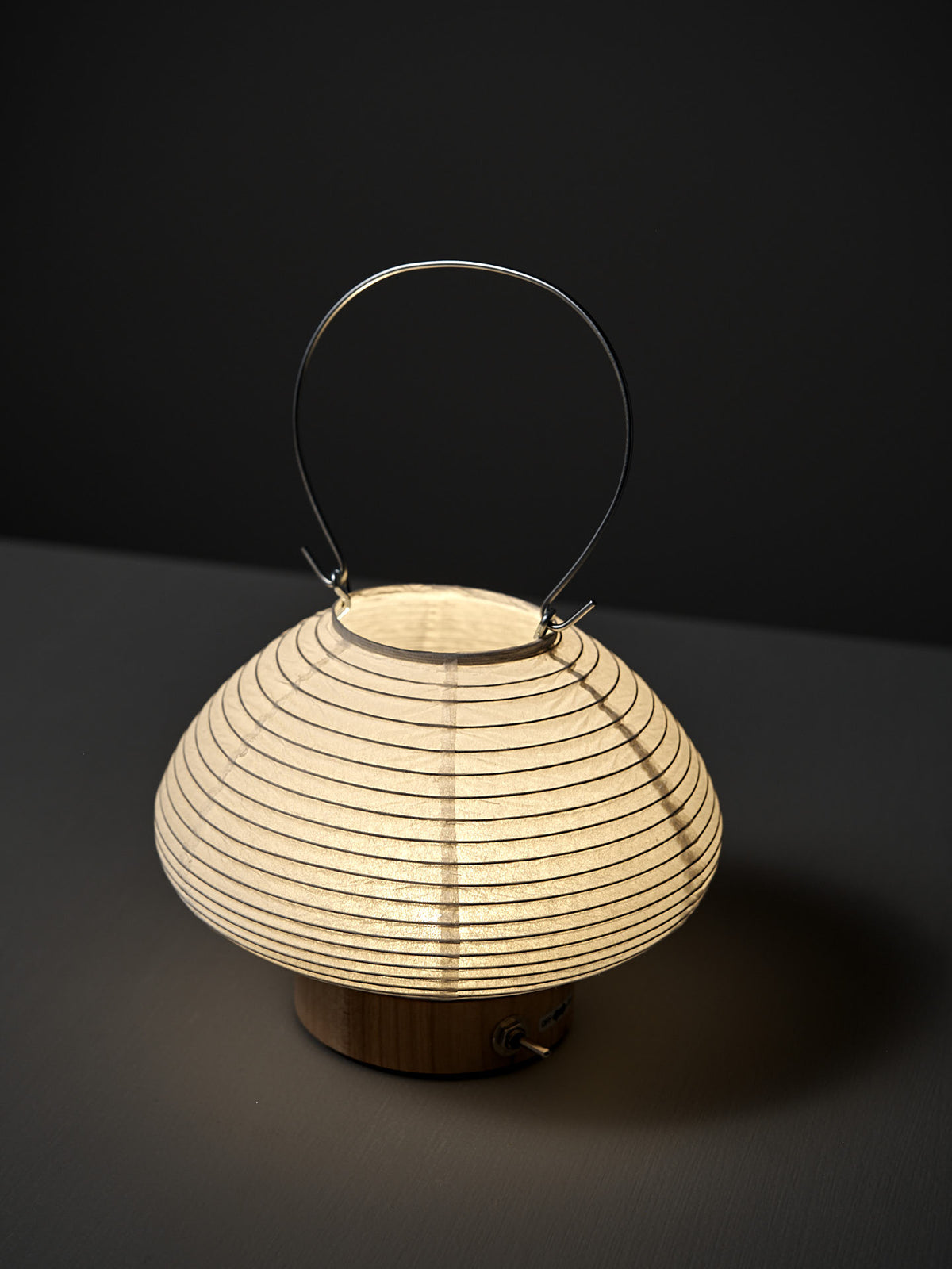 The Washi Paper LED Lantern – Musubi, designed by Rina Ono, features a round, ribbed washi paper design and metal handle, displayed on a dark surface with a dark background.
