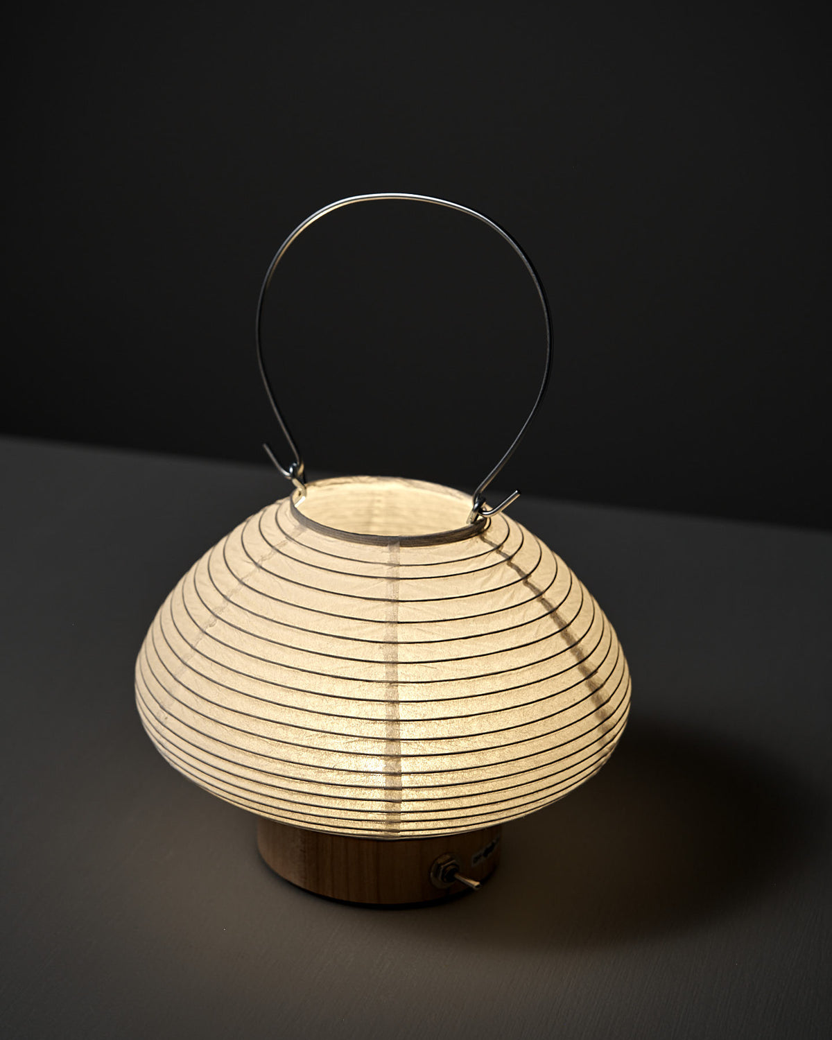 The Washi Paper LED Lantern – Musubi, designed by Rina Ono, features a round, ribbed washi paper design and metal handle, displayed on a dark surface with a dark background.