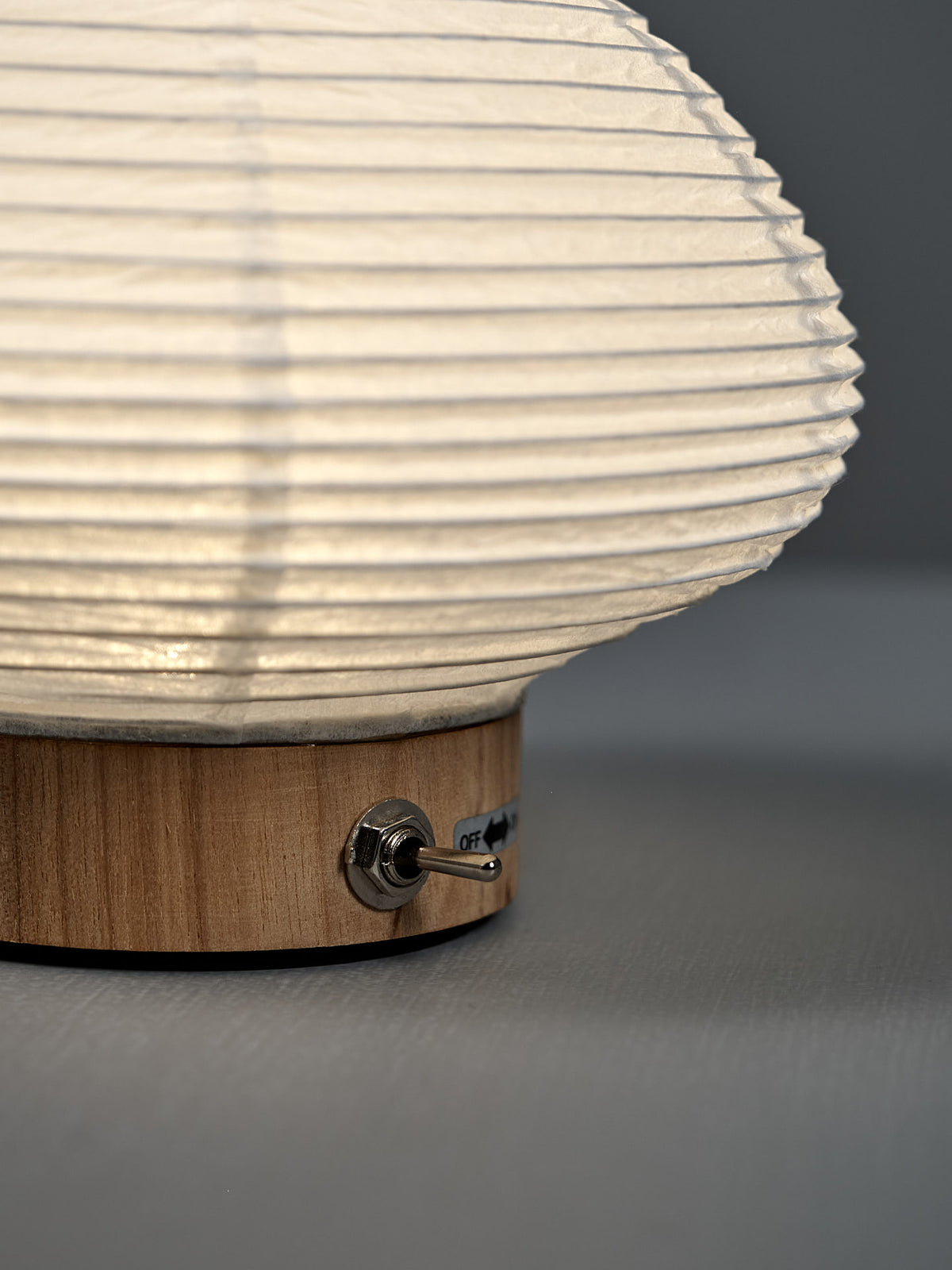 A close-up of the Washi Paper LED Lantern – Musubi, featuring a wooden base, white textured shade, and metal toggle switch labeled “off,” crafted by Japanese designer Rina Ono for Rinao design studio.